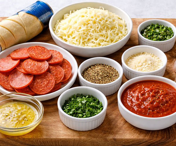 Ingredients for Mozzarella Pepperoni Croissant Rolls arranged on a wooden tray, including croissant dough, shredded mozzarella, pepperoni, butter, herbs, Parmesan, and marinara sauce.