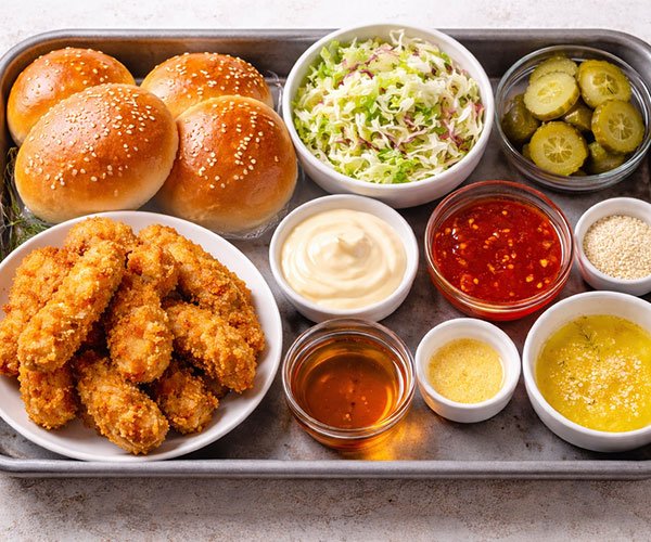 Ingredients for Bang Bang chicken sliders arranged on a tray, including round slider buns, breaded chicken tenders, sauces, shredded cabbage, pickles, butter, and sesame seeds.