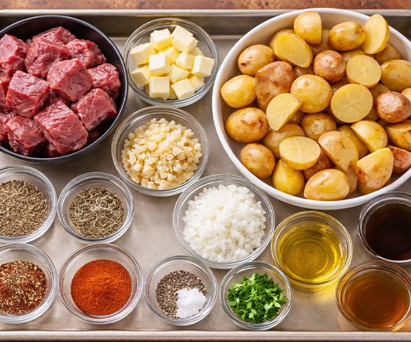 Tray with raw beef chunks, halved baby potatoes, butter cubes, garlic, onion, herbs, spices, oil, broth, and parsley neatly arranged.
