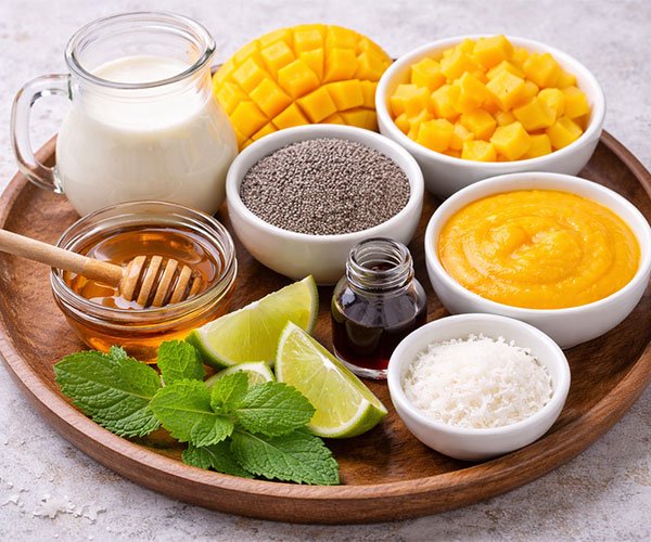 Coconut milk, chia seeds, mango, honey, vanilla, coconut, and mint arranged on a tray.