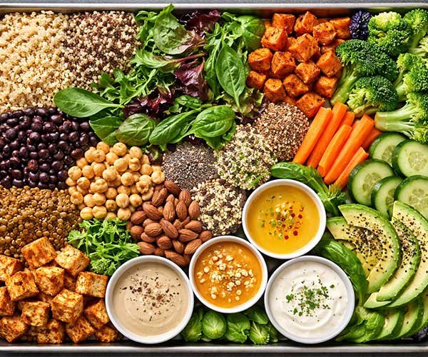 Tray filled with vegan Buddha bowl ingredients including grains, beans, tofu, vegetables, avocado, nuts, seeds, and creamy dressings arranged neatly.